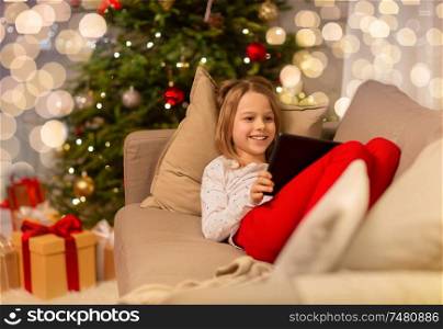 christmas, holidays and childhood concept - smiling girl with tablet pc computer lying on sofa at home. smiling girl with tablet pc at christmas home