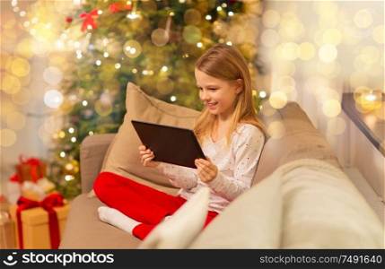 christmas, holidays and childhood concept - smiling girl with tablet pc computer sitting on sofa at home. smiling girl with tablet pc at christmas home
