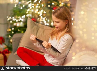 christmas, holidays and childhood concept - smiling girl with tablet pc computer sitting on sofa at home. smiling girl with tablet pc at christmas home