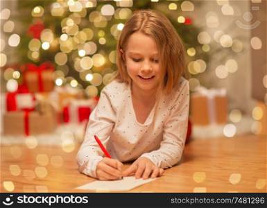 christmas, holidays and childhood concept - smiling girl writing wish list or letter to santa at home. smiling girl writing christmas wish list at home