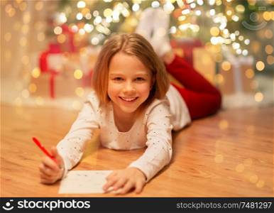 christmas, holidays and childhood concept - smiling girl writing wish list or letter to santa at home. smiling girl writing christmas wish list at home