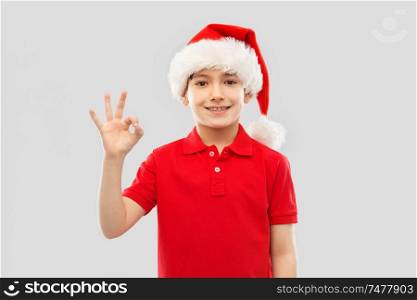 christmas, holidays and childhood concept - smiling little boy in red polo t-shirt and santa helper hat showing ok hand sign over grey background. smiling boy in santa helper hat showing ok gesture