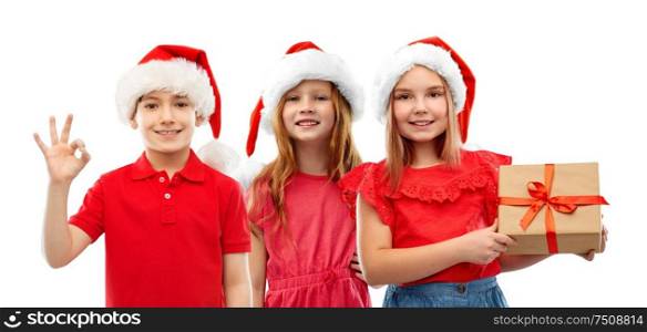 christmas, holidays and childhood concept - smiling little children in santa helper hats with gift box showing ok hand sign over white background. children in santa helper hats with christmas gift