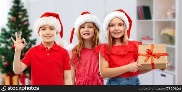 christmas, holidays and childhood concept - smiling little children in santa helper hats with gift box showing ok hand sign over home background. children in santa helper hats with christmas gift