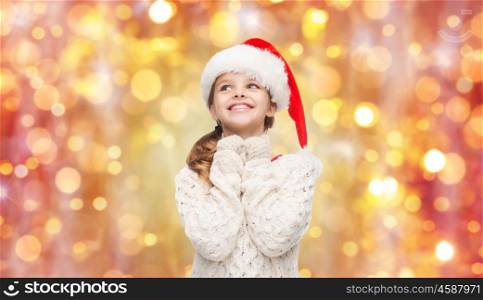 christmas, holidays and children concept - dreaming girl in santa helper hat over lights background