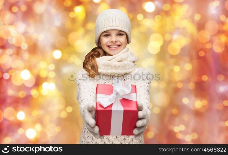 christmas, holidays and children concept -happy girl in hat, scarf and gloves with gift box over lights background