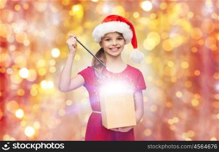 christmas, holidays and children concept - smiling girl in santa helper hat with gift box and magic wand over lights background