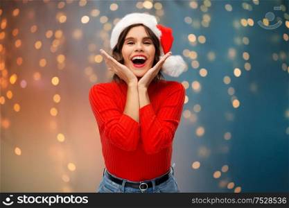 christmas, holidays and emotions concept - happy smiling young woman in santa helper hat over festive lights background. happy young woman in santa hat on christmas