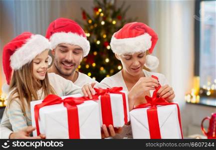 christmas, holidays and family concept - happy mother, father and daughter in santa hats with gifts sitting on couch at home. happy family with christmas gifts at home