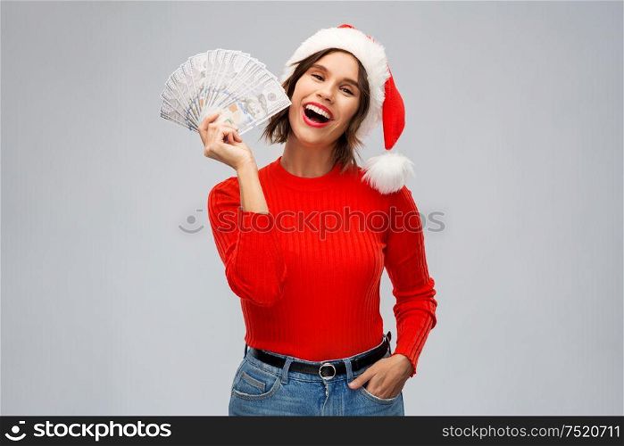christmas, holidays and finance concept - happy smiling young woman in santa helper hat holding dollar money banknotes over grey background. happy woman in santa hat with money on christmas