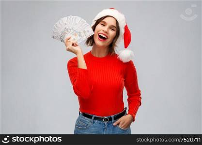 christmas, holidays and finance concept - happy smiling young woman in santa helper hat holding dollar money banknotes over grey background. happy woman in santa hat with money on christmas