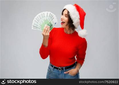 christmas, holidays and finance concept - happy smiling young woman in santa helper hat holding euro money banknotes over grey background. happy woman in santa hat with money on christmas