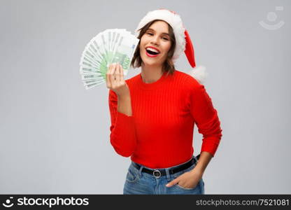 christmas, holidays and finance concept - happy smiling young woman in santa helper hat holding euro money banknotes over grey background. happy woman in santa hat with money on christmas