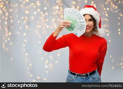 christmas, holidays and finance concept - happy smiling young woman in santa helper hat holding euro money banknotes over festive lights on grey background. happy woman in santa hat with money on christmas