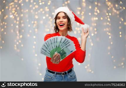 christmas, holidays and finance concept - happy smiling young woman in santa helper hat holding euro money banknotes over festive lights on grey background. happy woman in santa hat with money on christmas