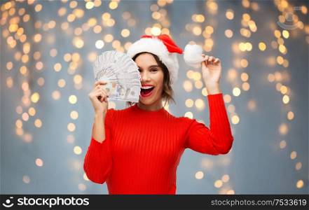 christmas, holidays and finance concept - happy smiling young woman in santa helper hat holding dollar money banknotes over festive lights on grey background. happy woman in santa hat with money on christmas