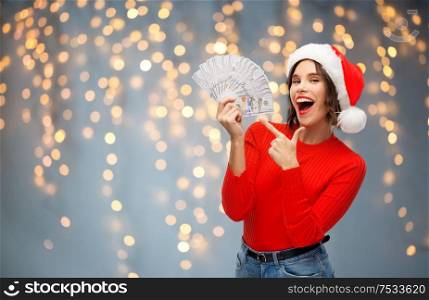christmas, holidays and finance concept - happy smiling young woman in santa helper hat holding dollar money banknotes over festive lights on grey background. happy woman in santa hat with money on christmas