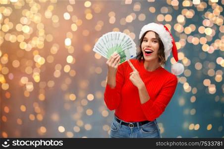 christmas, holidays and finance concept - happy smiling young woman in santa helper hat holding euro money banknotes over festive lights background. happy woman in santa hat with money on christmas