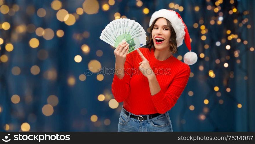christmas, holidays and finance concept - happy smiling young woman in santa helper hat holding euro money banknotes over festive lights on dark night background. happy woman in santa hat with money on christmas