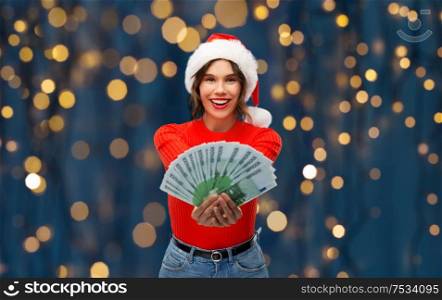 christmas, holidays and finance concept - happy smiling young woman in santa helper hat holding euro money banknotes over festive lights on dark night background. happy woman in santa hat with money on christmas