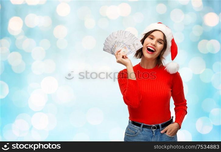 christmas, holidays and finance concept - happy smiling young woman in santa helper hat holding dollar money banknotes over festive lights on blue background. happy woman in santa hat with money on christmas