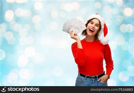 christmas, holidays and finance concept - happy smiling young woman in santa helper hat holding dollar money banknotes over festive lights on blue background. happy woman in santa hat with money on christmas