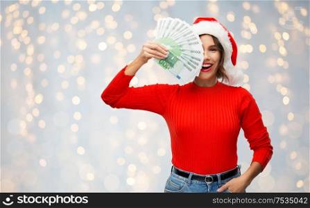 christmas, holidays and finance concept - happy smiling young woman in santa helper hat holding euro money banknotes over festive lights background. happy woman in santa hat with money on christmas