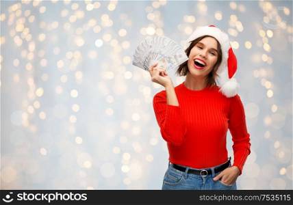christmas, holidays and finance concept - happy smiling young woman in santa helper hat holding dollar money banknotes over festive lights background. happy woman in santa hat with money on christmas
