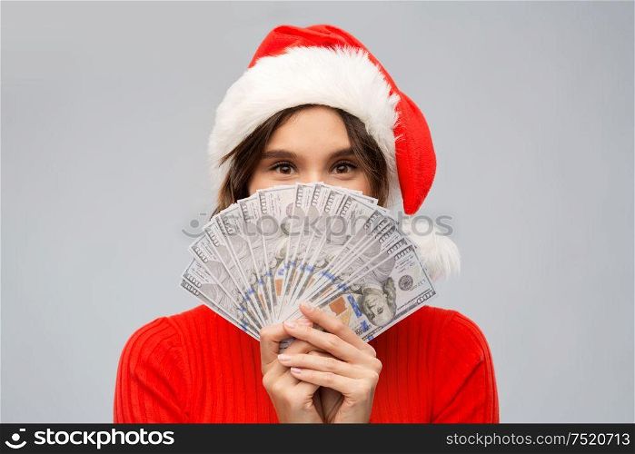 christmas, holidays and finance concept - happy young woman in santa helper hat hiding behind dollar money banknotes over grey background. happy woman in santa hat with money on christmas