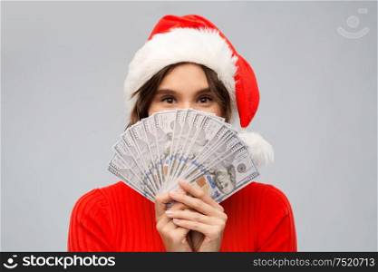 christmas, holidays and finance concept - happy young woman in santa helper hat hiding behind dollar money banknotes over grey background. happy woman in santa hat with money on christmas
