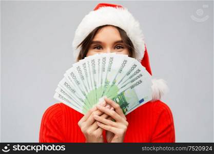 christmas, holidays and finance concept - happy young woman in santa helper hat hiding behind euro money banknotes over grey background. happy woman in santa hat with money on christmas
