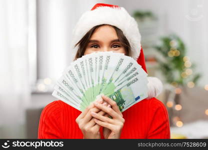 christmas, holidays and finance concept - happy young woman in santa helper hat hiding behind euro money banknotes over grey background. happy woman in santa hat with money on christmas