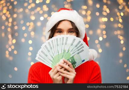 christmas, holidays and finance concept - happy young woman in santa helper hat hiding behind euro money banknotes over festive lights on grey background. happy woman in santa hat with money on christmas