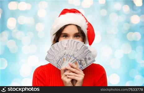christmas, holidays and finance concept - happy young woman in santa helper hat hiding behind dollar money banknotes over festive lights on blue background. happy woman in santa hat with money on christmas
