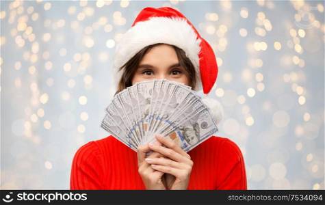 christmas, holidays and finance concept - happy young woman in santa helper hat hiding behind dollar money banknotes over festive lights background. happy woman in santa hat with money on christmas