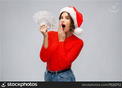 christmas, holidays and finance concept - surprised young woman in santa helper hat holding dollar money banknotes over grey background. surprised woman in santa hat with money