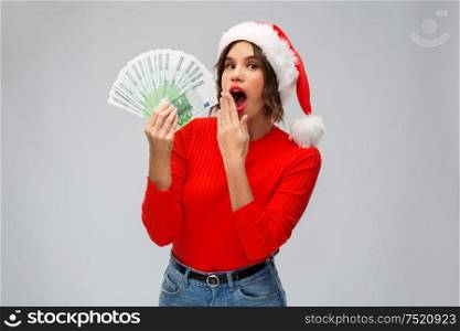 christmas, holidays and finance concept - surprised young woman in santa helper hat holding euro money banknotes over grey background. surprised woman in santa hat with money