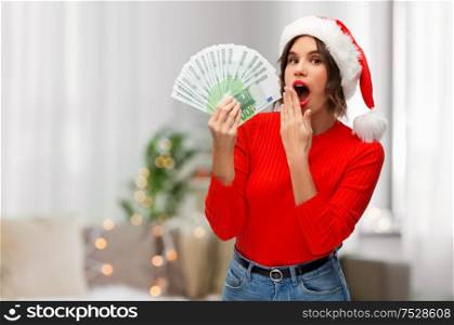 christmas, holidays and finance concept - surprised young woman in santa helper hat holding euro money banknotes over grey background. woman in santa hat with money on christmas
