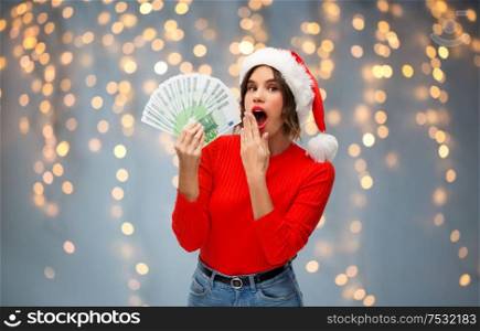 christmas, holidays and finance concept - surprised young woman in santa helper hat holding euro money banknotes over festive lights on grey background. woman in santa hat with money on christmas