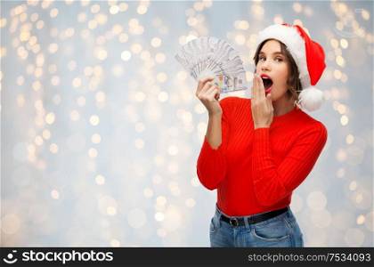 christmas, holidays and finance concept - surprised young woman in santa helper hat holding dollar money banknotes over festive lights background. surprised woman in santa hat with money