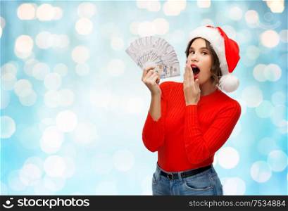 christmas, holidays and finance concept - surprised young woman in santa helper hat holding dollar money banknotes over festive lights on blue background. surprised woman in santa hat with money