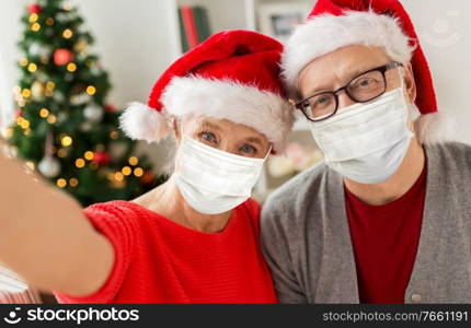 christmas, holidays and health concept - happy smiling senior couple wearing face protective medical mask for protection from virus disease in santa hats taking selfie at home. senior couple in masks taking christmas selfie