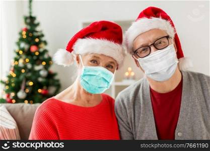 christmas, holidays and health concept - senior couple in santa hats wearing face protective medical mask for protection from virus disease at home. senior couple in masks and santa hats on christmas
