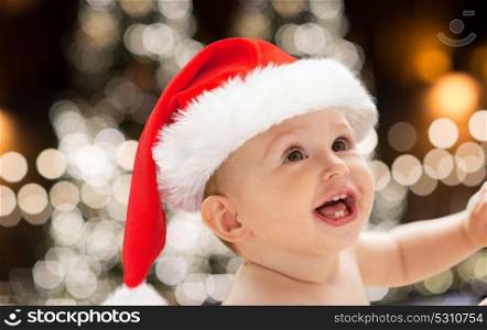 christmas, holidays and people concept - close up of happy little baby boy or girl in santa hat over lights background. close up of little baby in santa hat at christmas