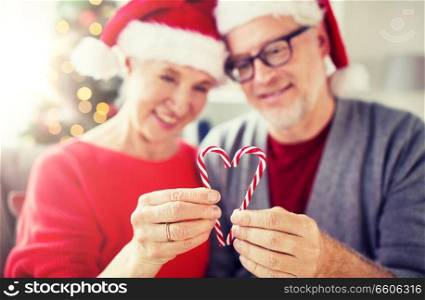 christmas, holidays and people concept - close up of happy smiling senior couple in santa hats with heart of candy canes at home. senior couple with heart of christmas candy canes