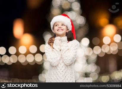 christmas, holidays and people concept - dreaming girl in santa helper hat over lights background. dreaming girl in santa helper hat