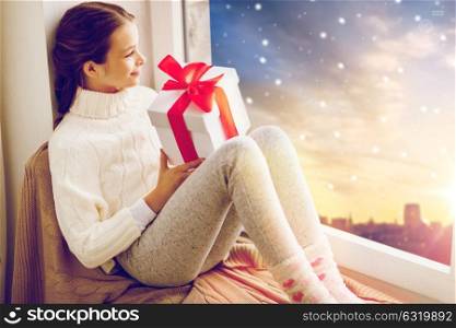 christmas, holidays and people concept - happy beautiful girl with gift sitting on window sill in winter at home over snow city background. girl with christmas gift on window sill in winter