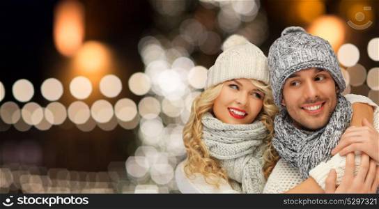 christmas, holidays and people concept - happy couple in hats and scarf hugging over night lights background. happy couple hugging over christmas lights