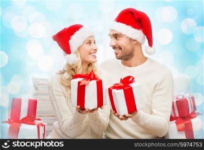 christmas, holidays and people concept - happy couple in santa hats exchanging gifts over blue holidays lights background