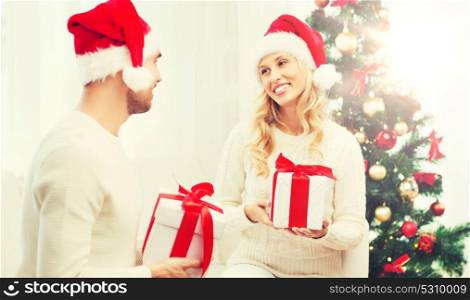 christmas, holidays and people concept - happy couple in santa hats exchanging gifts at home. happy couple at home exchanging christmas gifts